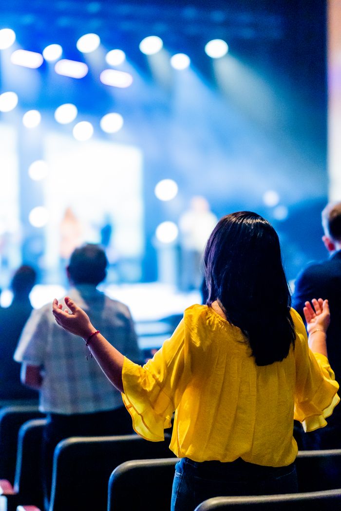 bedroom-bliss A woman in a yellow blouse with hands raised at a lively concert event indoors.